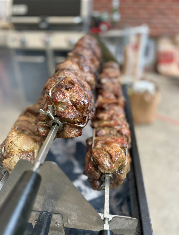 Skewered roasted meats on a rotisserie grill with a blurred outdoor greek kontosouvli kotosouvli octagrills pork charcoal background