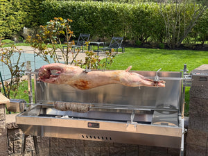 Outdoor setting with a large grill on a patio, featuring a whole lamb being roasted on a spit.  Seen here with our optional add on skewer kit spinning a kokoretsi on a souvla.