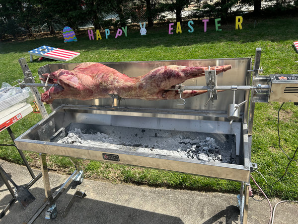 Barbecue rotisserie spit with a large lamb spinning, set against a grassy area with 'Happy Easter' decorations. Greek easter celebraton